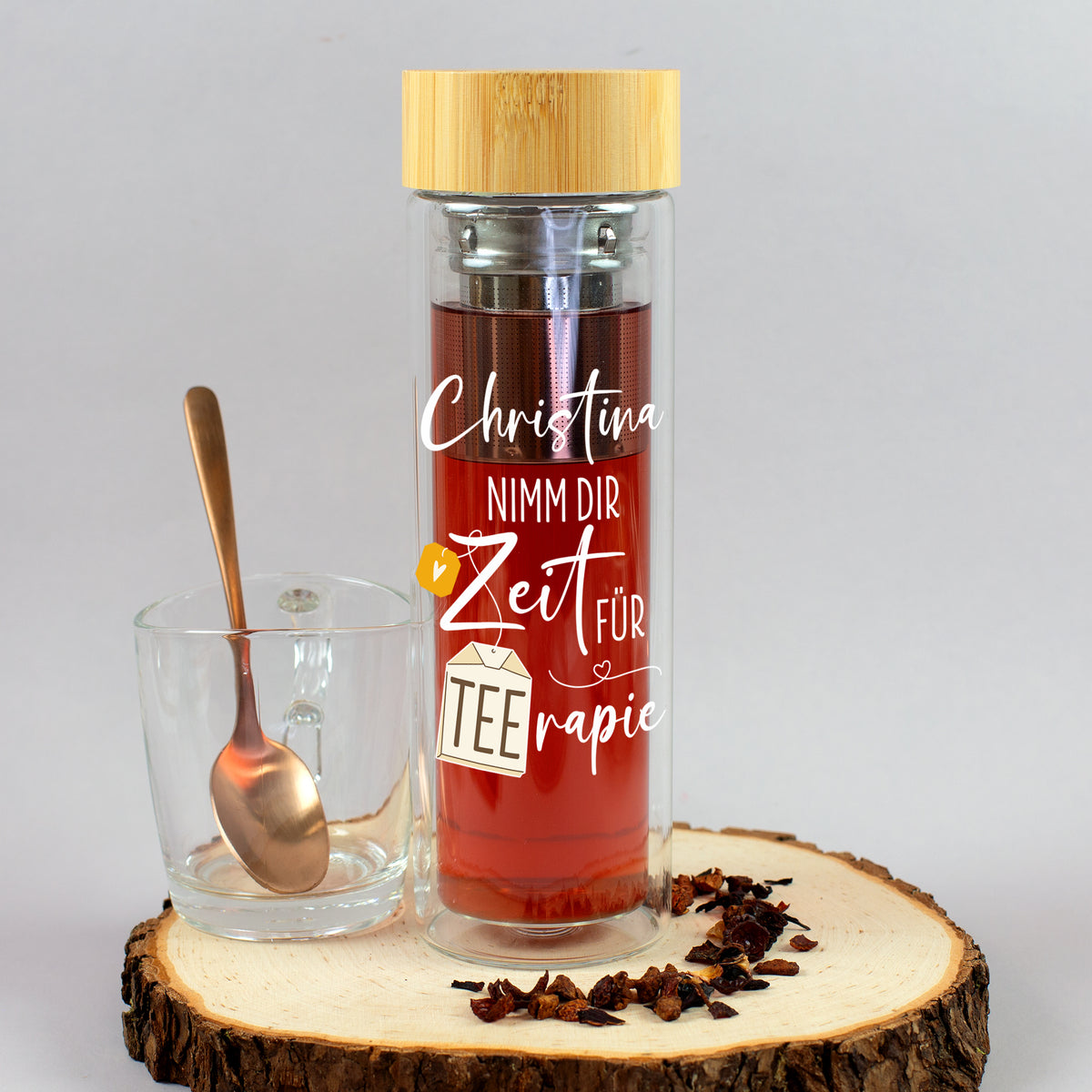 Personalisierte Teeflasche mit rotem Tee, Edelstahl-Teesieb, Bambusdeckel und Wunschname Christina neben Teeglas und losem Tee auf Holzscheibe