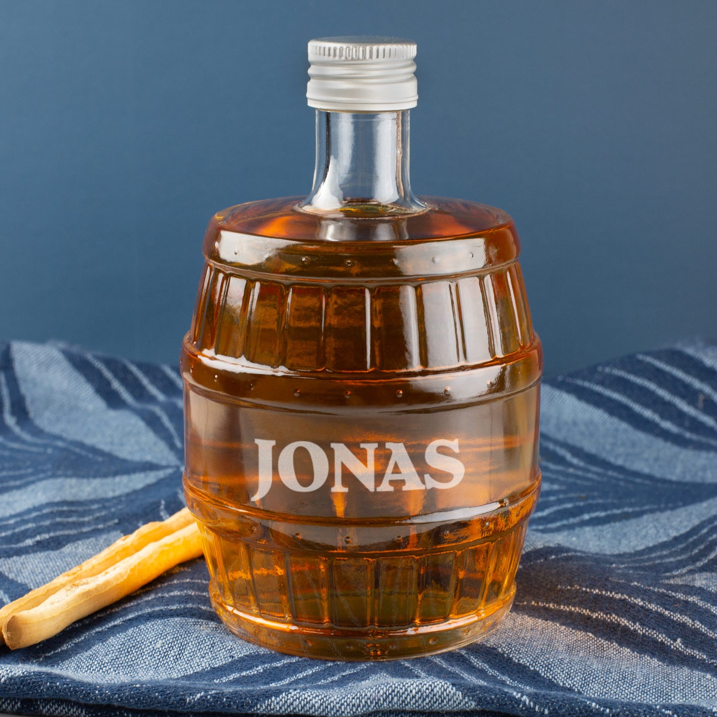 Gravierte Whisky-Flasche in Fassform mit Name JONAS, dekoriert mit Salzstange, auf blauem Tuch