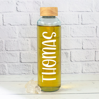 Personalisierte Glasflasche mit weißem Namensaufdruck Thomas, gefüllt mit gelbem Getränk, mit Holzdeckel vor heller Steinwand