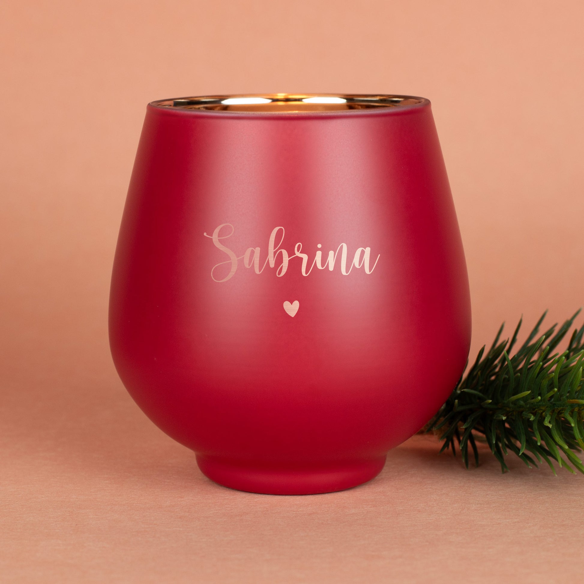 Rotes Windlicht mit Gravur „Sabrina“ und Herzmotiv, auf beigem Untergrund mit Tannenzweig dekoriert.