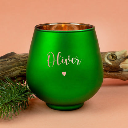 Grünes Windlicht mit Gravur „Oliver“ und Herzmotiv, dekorativ vor Holzstück und Tannenzweig auf orangefarbenem Untergrund.