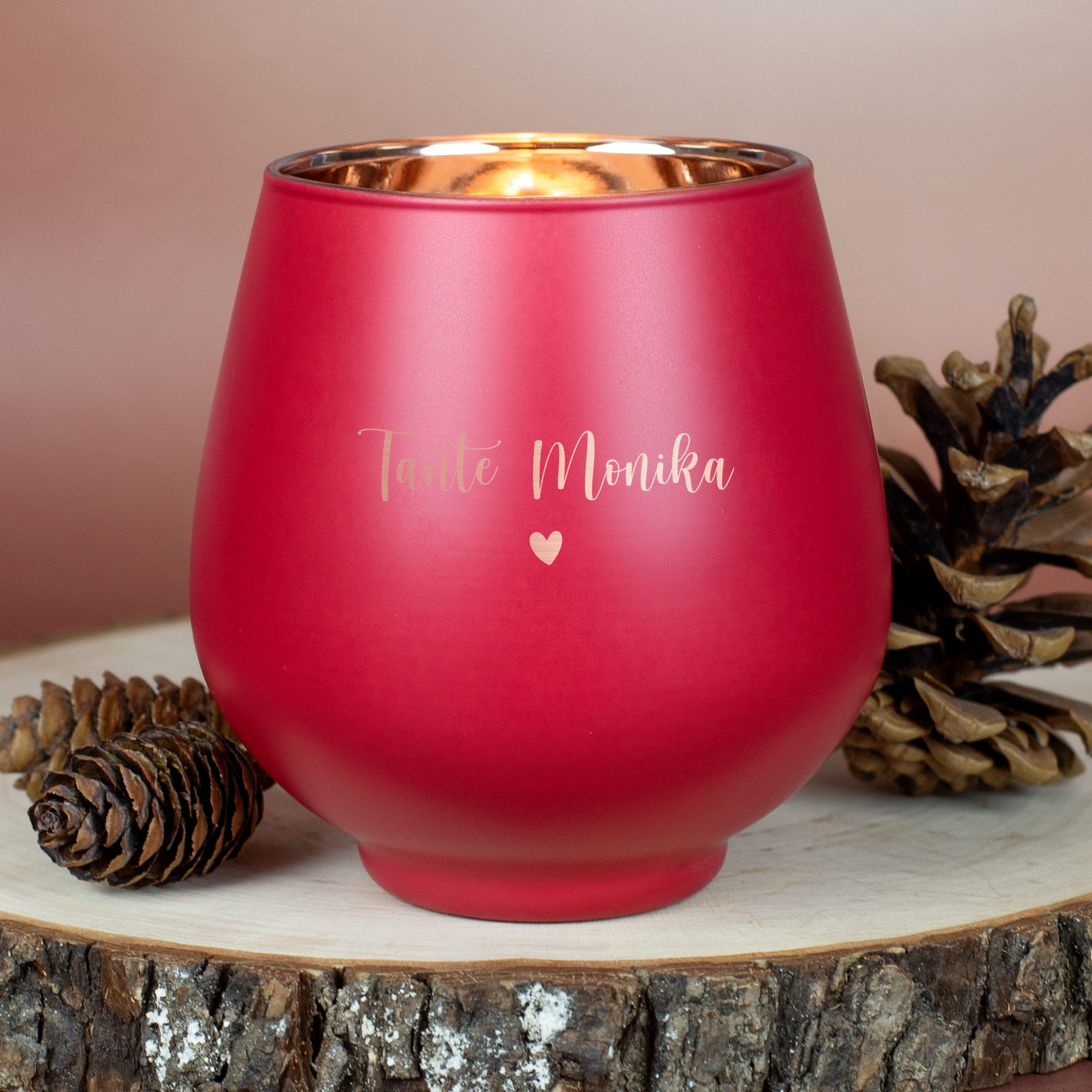 Rotes Windlicht mit Gravur „Tante Monika“ und Herzsymbol, auf Holzscheibe mit Kiefernzapfen arrangiert.