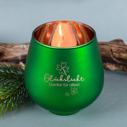 Grünes Windlicht mit Gravur „Glückslicht Danke für alles!“, vor Holzstück und Tannenzweig arrangiert
