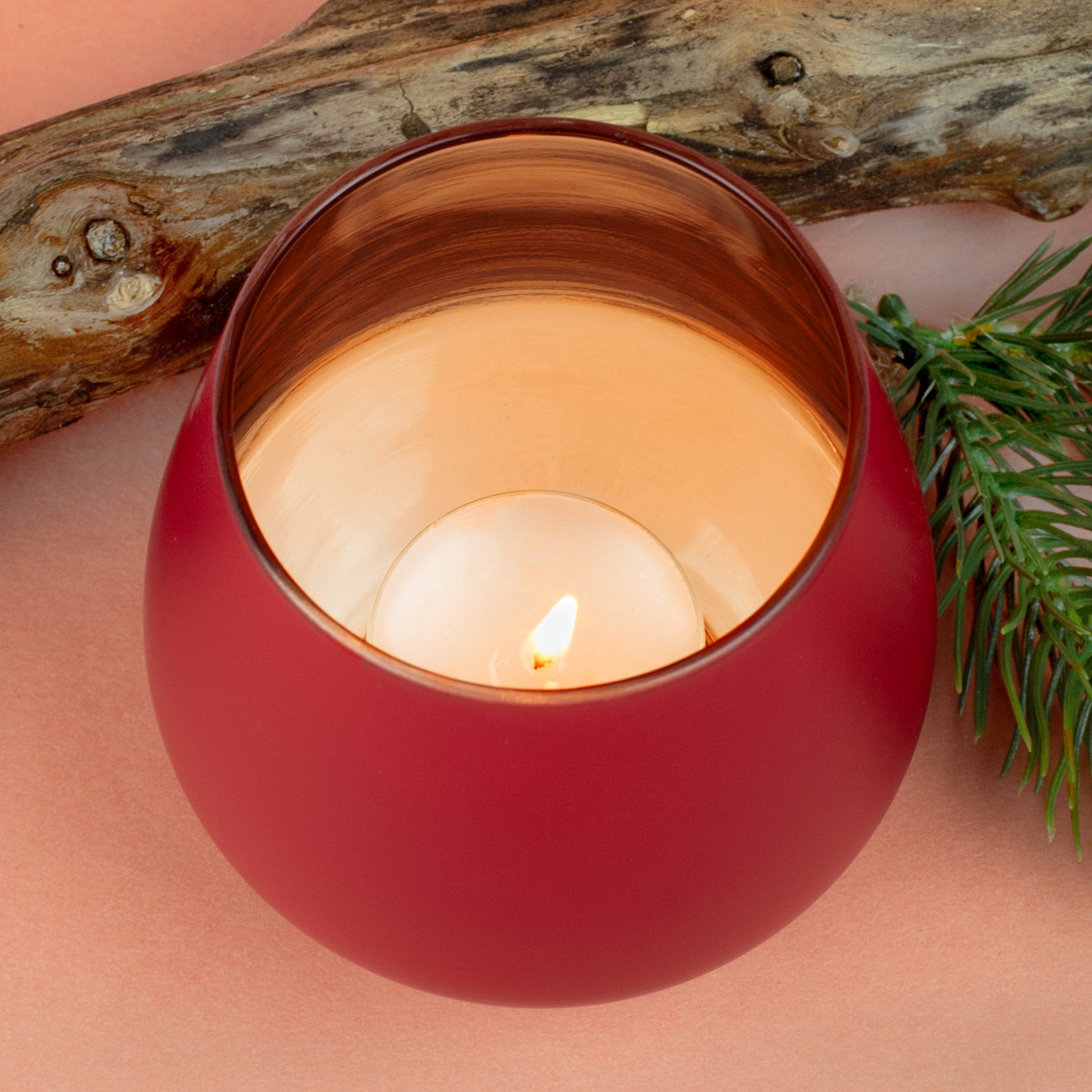Detailaufnahme eines brennenden Teelichts im roten Windlicht mit kupferfarbener Innenfläche, umgeben von Holz und Tannenzweig.