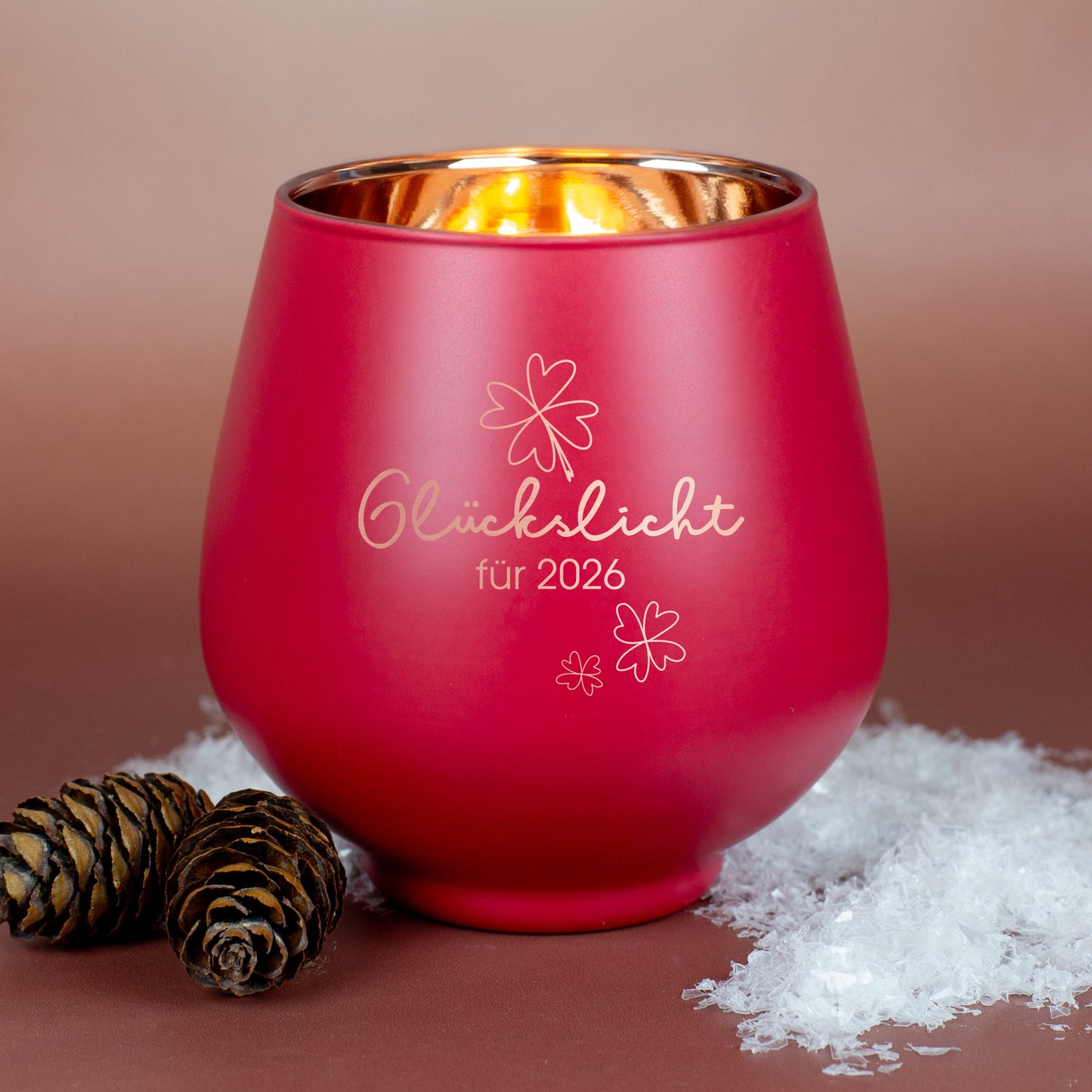 Rotes Windlicht mit Gravur „Glückslicht für 2026“, auf braunem Hintergrund mit Zapfen und Kunstschnee