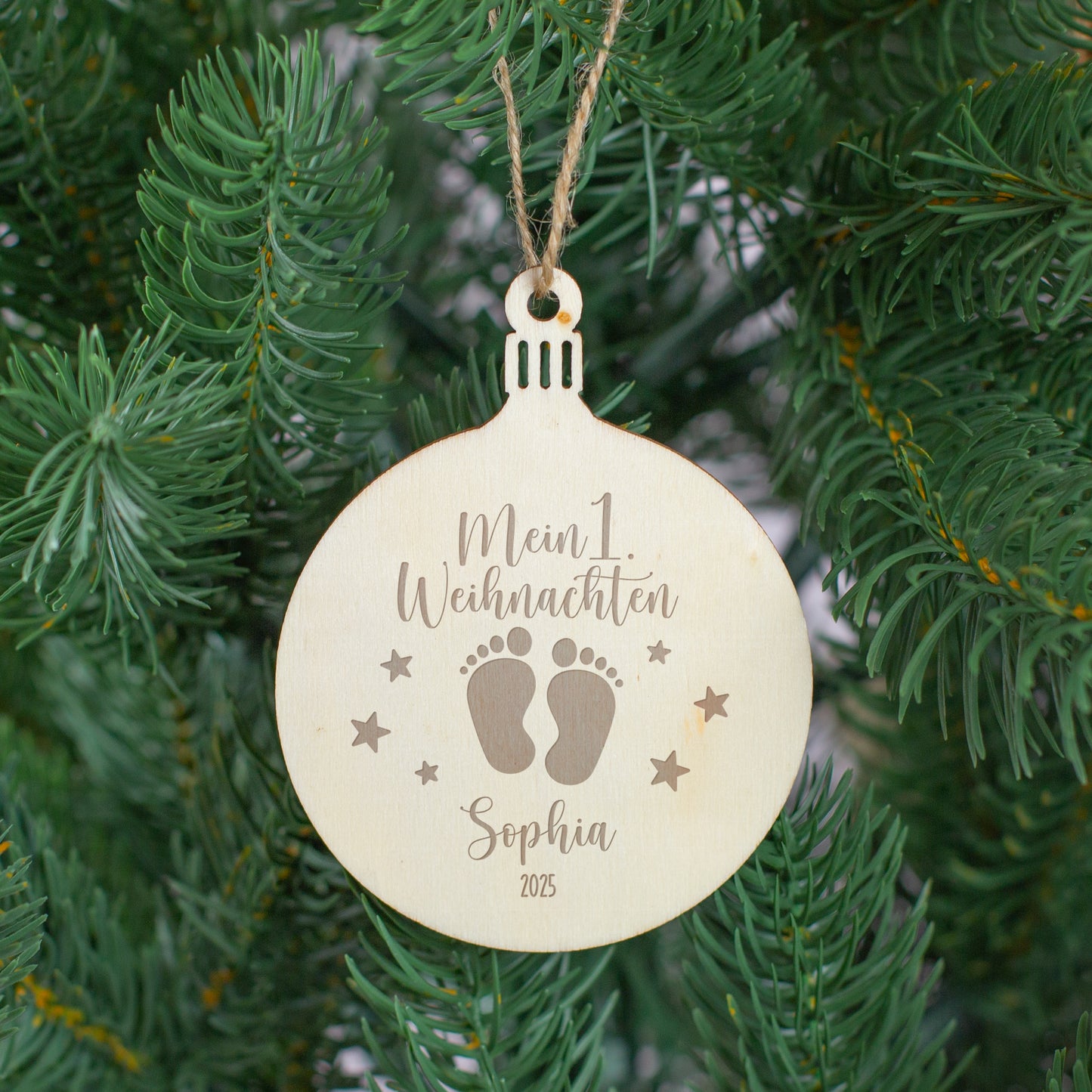 Personalisierter Holzanhänger „Mein 1. Weihnachten“ mit Gravur und Juteschnur, hängend am Weihnachtsbaum zwischen grünen Tannenzweigen.