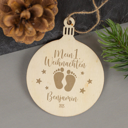 Weihnachtsanhänger aus Holz mit Babyfüßchen-Motiv und Gravur „Mein 1. Weihnachten – Benjamin 2025“, liegend neben Zapfen und Zweig.