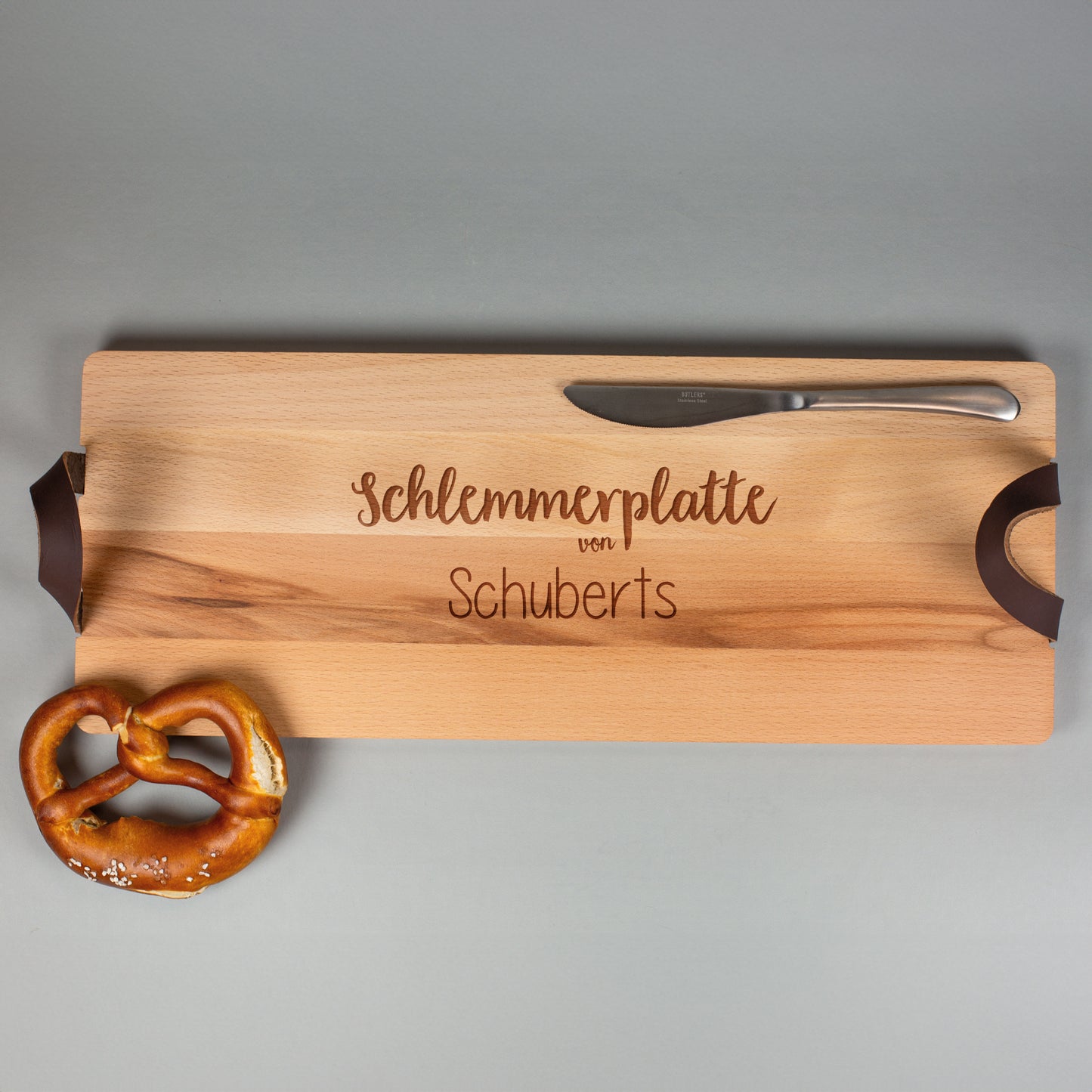 Servierbrett „Schlemmerplatte von Schuberts“ aus Buchenholz mit Ledergriffen, frontal fotografiert mit Brezel und Messer.