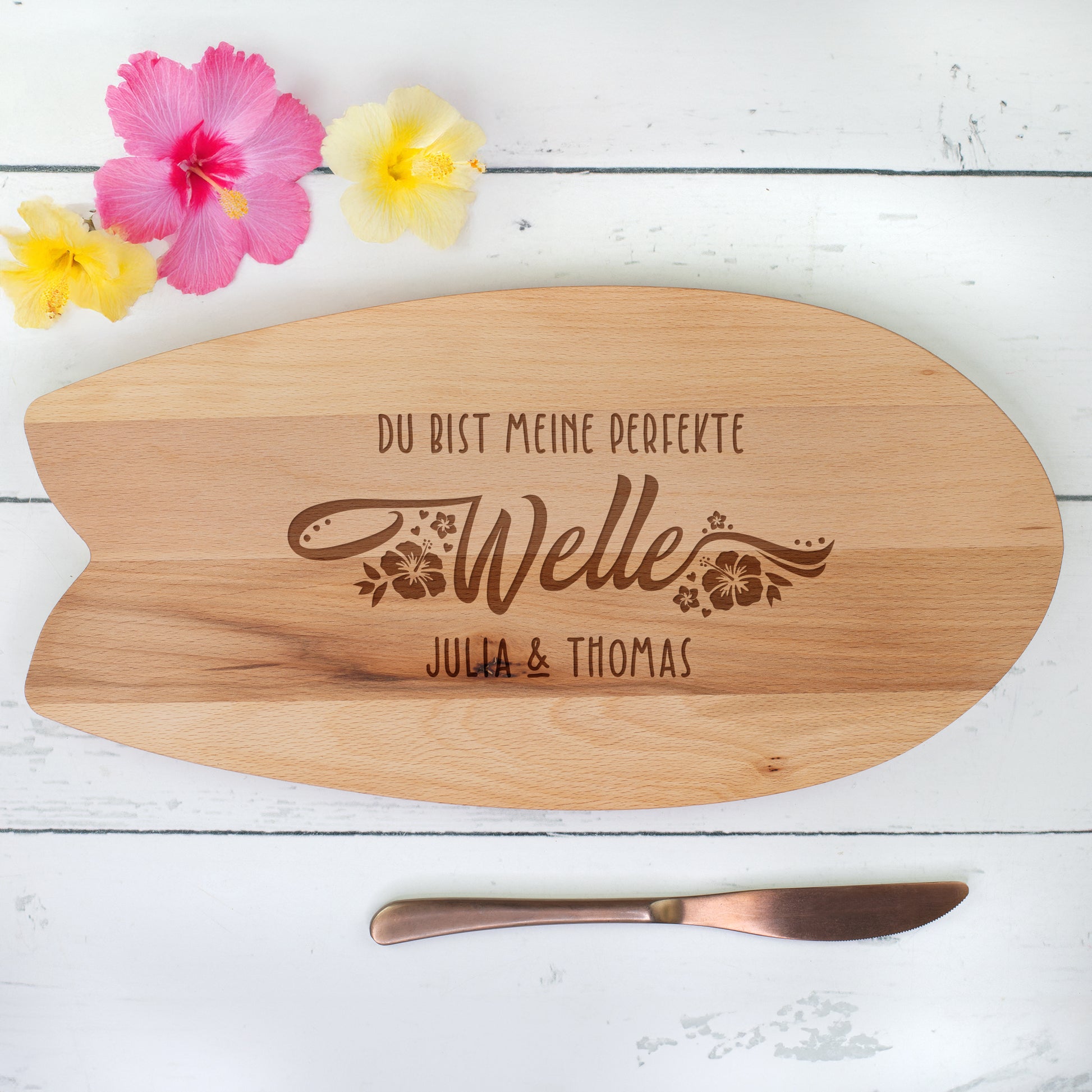 Personalisiertes Servierbrett im Surfbrett-Design aus Holz mit Gravur „Du bist meine perfekte Welle“ und Namen – Draufsicht auf weißem Holzhintergrund mit Blumen