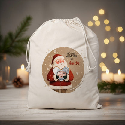 Großer naturfarbener Geschenksack mit Weihnachtsmann-Motiv, personalisiert mit Foto im Herz und Text „Fröhliche Weihnachten für Amelie“, auf weihnachtlich dekoriertem Tisch mit Kerzen im Hintergrund.