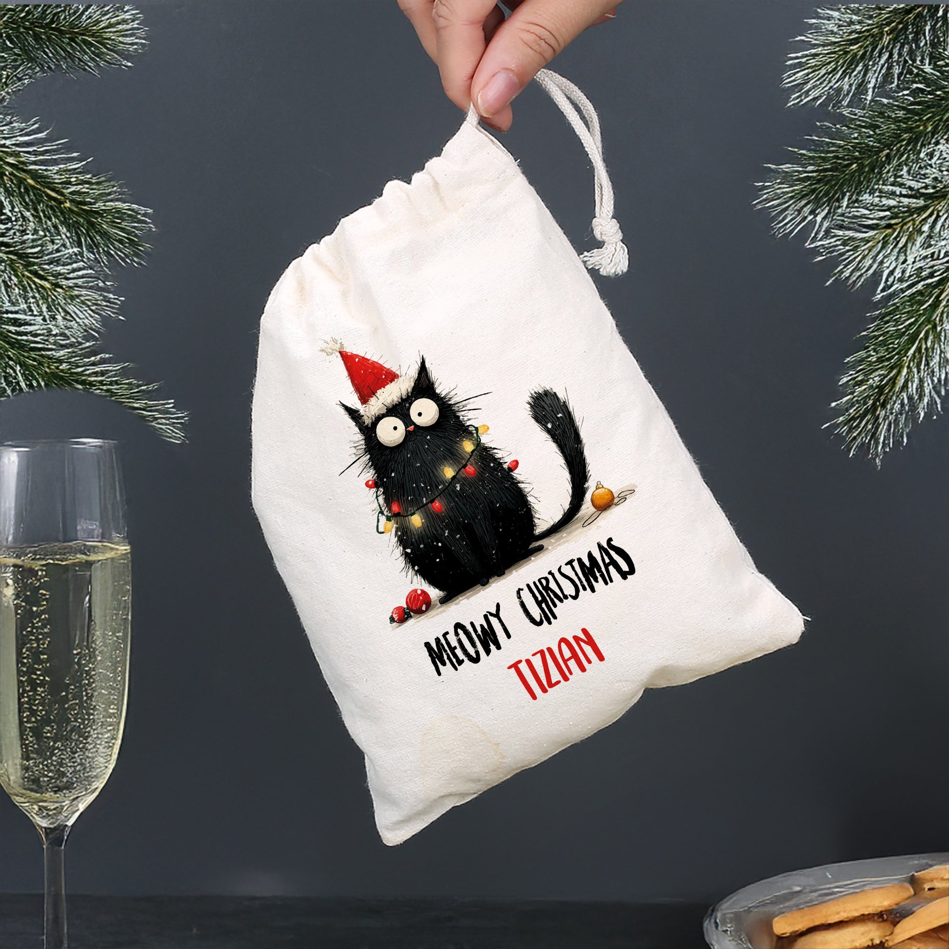 Personalisierter Baumwoll-Geschenksack mit schwarzer Weihnachtskatze und Aufschrift „Meowy Christmas – Tizian“, Sektglas im Hintergrund