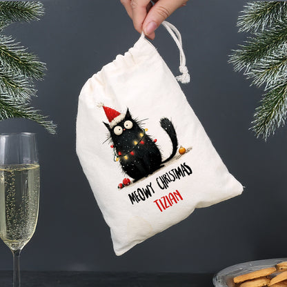 Personalisierter Baumwoll-Geschenksack mit schwarzer Weihnachtskatze und Aufschrift „Meowy Christmas – Tizian“, Sektglas im Hintergrund