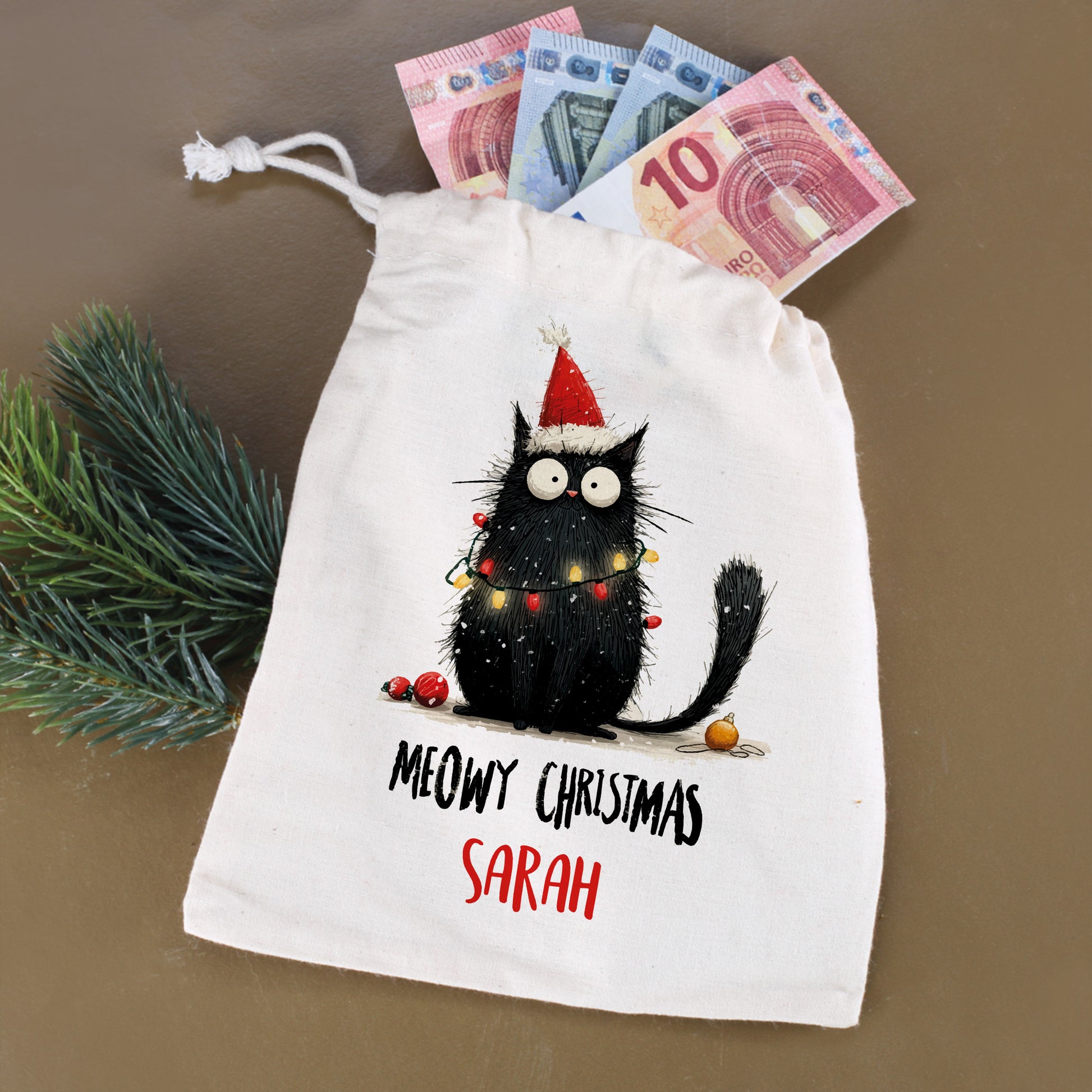 Stoffbeutel mit Katzenmotiv „Meowy Christmas – Sarah“, befüllt mit Geldscheinen und Tannenzweig daneben
