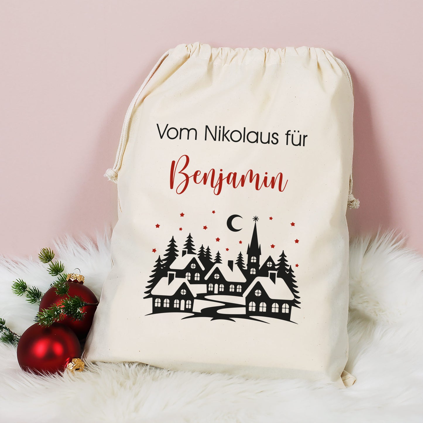 Mittlerer Baumwoll-Geschenksack mit Aufdruck „Vom Nikolaus für Benjamin“ und Winterlandschaft, dekoriert mit Christbaumkugeln auf weißem Fell.