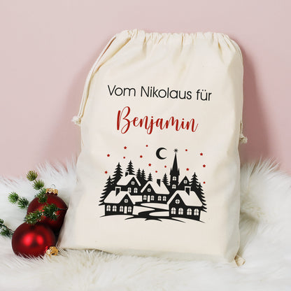 Mittlerer Baumwoll-Geschenksack mit Aufdruck „Vom Nikolaus für Benjamin“ und Winterlandschaft, dekoriert mit Christbaumkugeln auf weißem Fell.