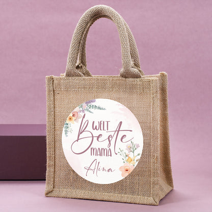 Jute-Geschenktasche „Weltbeste Mama“ mit floralem Motiv & personalisierbarem Namen seitlich stehend vor rosa Hintergrund