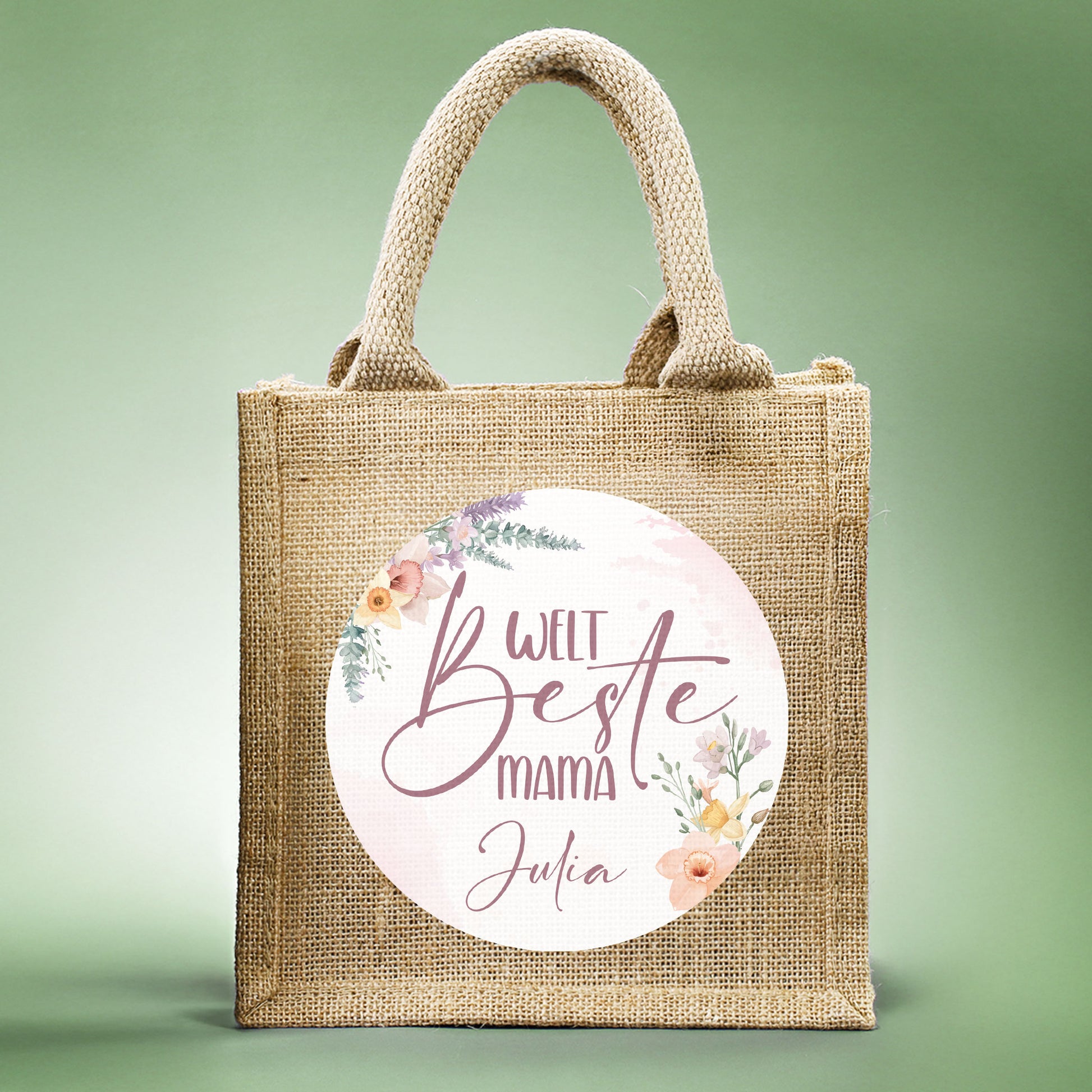 Jute-Geschenktasche „Weltbeste Mama“ mit floralem Motiv & personalisierbarem Namen frontal fotografiert vor grünem Hintergrund