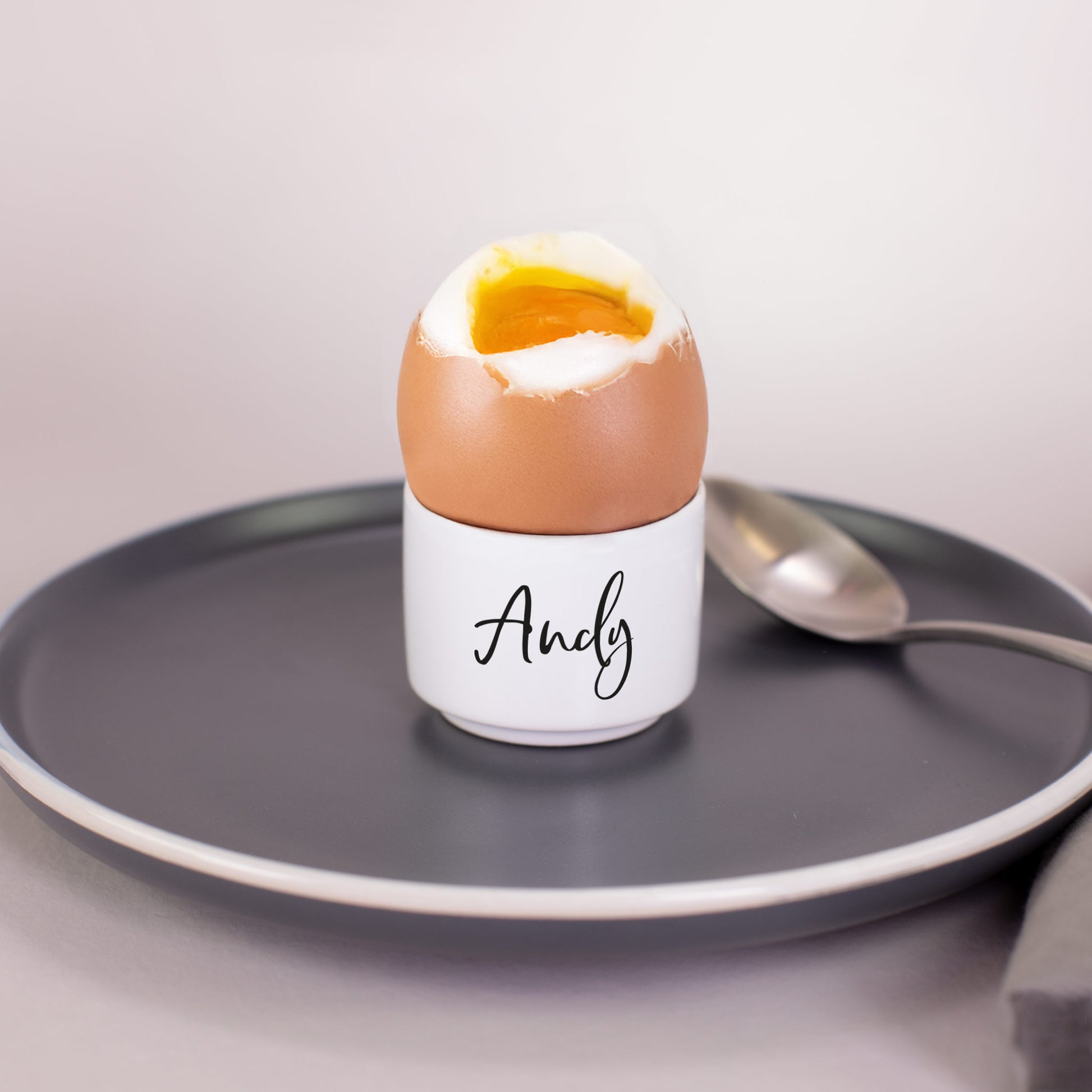 Gekochtes Ei in weißem Keramik-Eierbecher mit schwarzem Namensaufdruck „Andy“ auf grauem Teller mit Löffel
