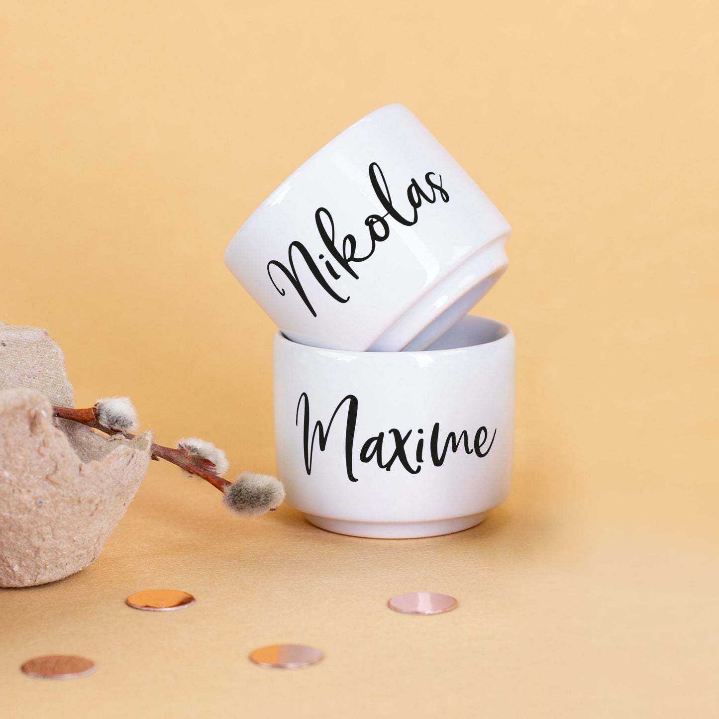 Zwei weiße Eierbecher mit den Namen „Nikolas“ und „Maxime“ auf gelbem Hintergrund, daneben Deko mit Weidenkätzchen