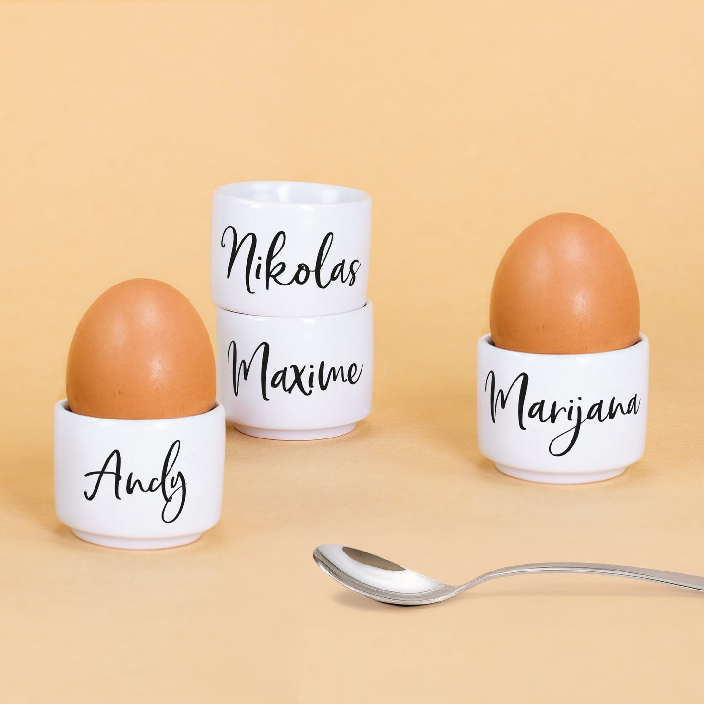 Vier weiße Keramik-Eierbecher mit Eiern und Namen „Andy“, „Marijana“, „Nikolas“ und „Maxime“ aufgelistet auf gelbem Hintergrund