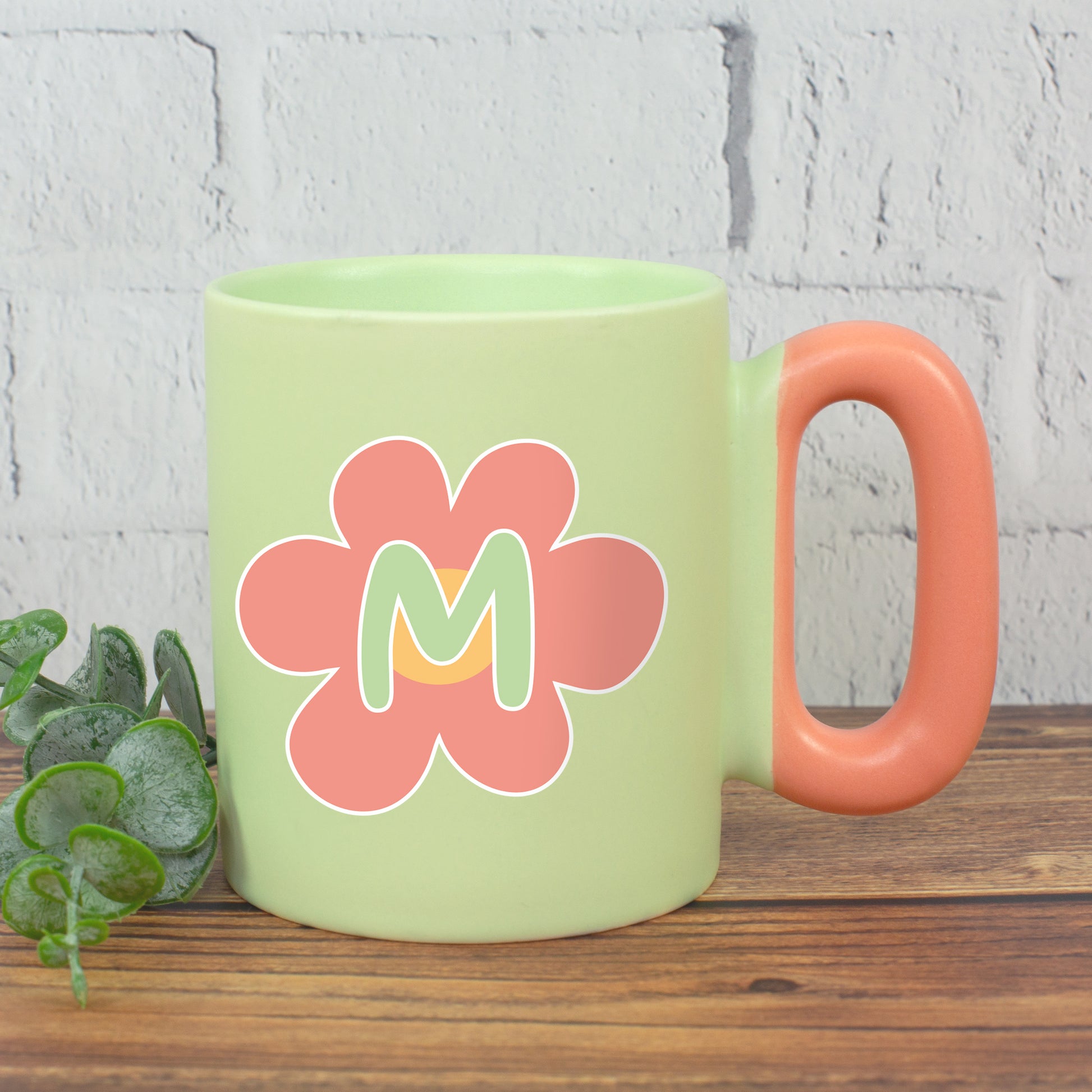 Grüne Tasse mit rosa Blume und Initiale M und farbigem Griff neben Pflanze