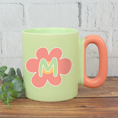 Grüne Tasse mit rosa Blume und Initiale M und farbigem Griff neben Pflanze