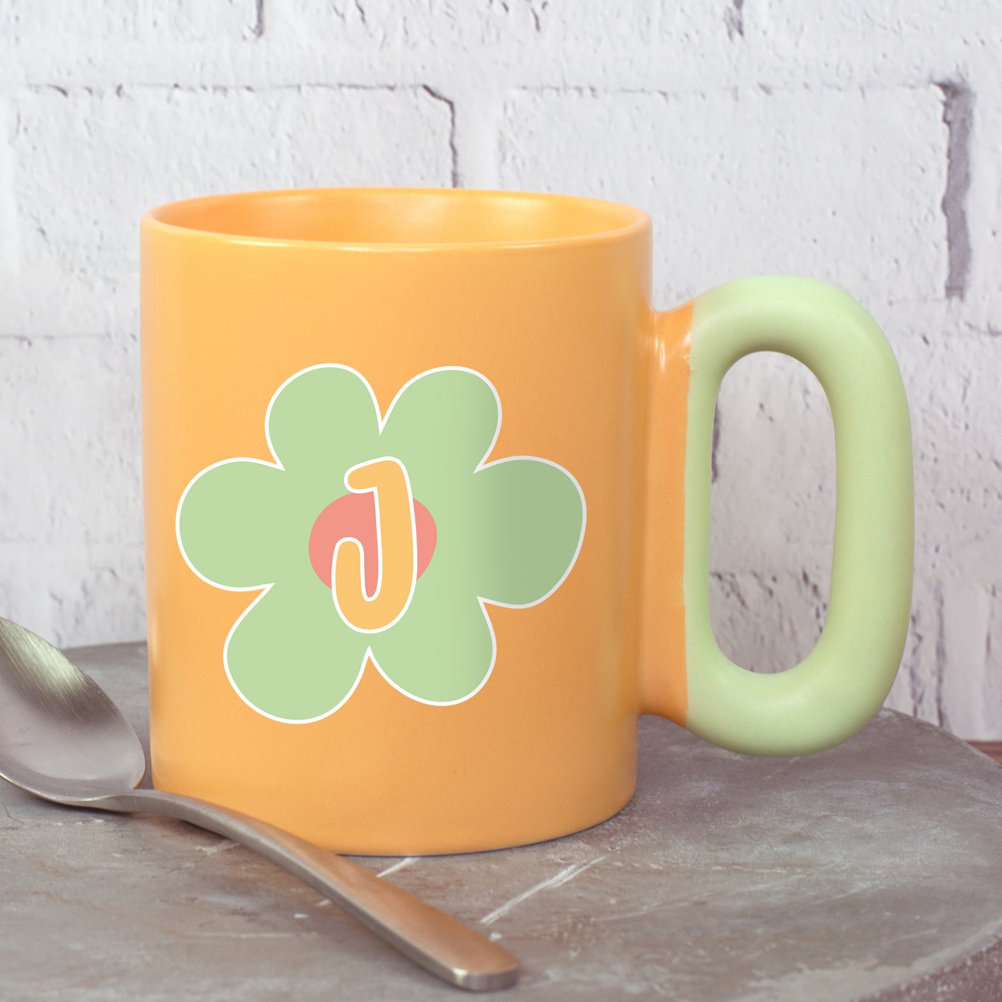 Orange Tasse mit grüner Blume und Initiale J mit ovalem Griff neben Löffel
