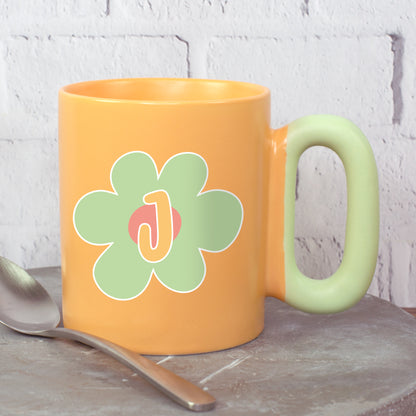 Orange Tasse mit grüner Blume und Initiale J mit ovalem Griff neben Löffel