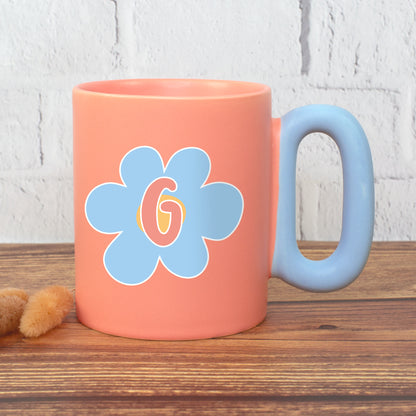 Korallfarbene Tasse mit blauer Blume und Initiale G auf Holzfläche