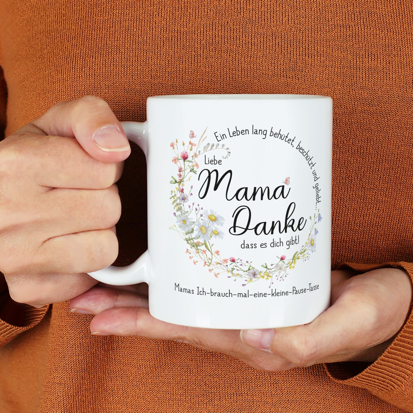Weiße Keramiktasse mit seidenglänzendem Finish, bedruckt mit einem Wiesenblumen-Kranz und dem Spruch „Ein Leben lang behütet, beschützt und geliebt…“. In der Mitte steht „Liebe Mama, danke, dass es dich gibt!“, ergänzt durch einen individuellen Wunschtext. Die Tasse wird von einer Person in einem warmen, orangefarbenen Pullover gehalten. Ein liebevolles Geschenk für Mütter zum Muttertag, Geburtstag oder als Dankeschön.