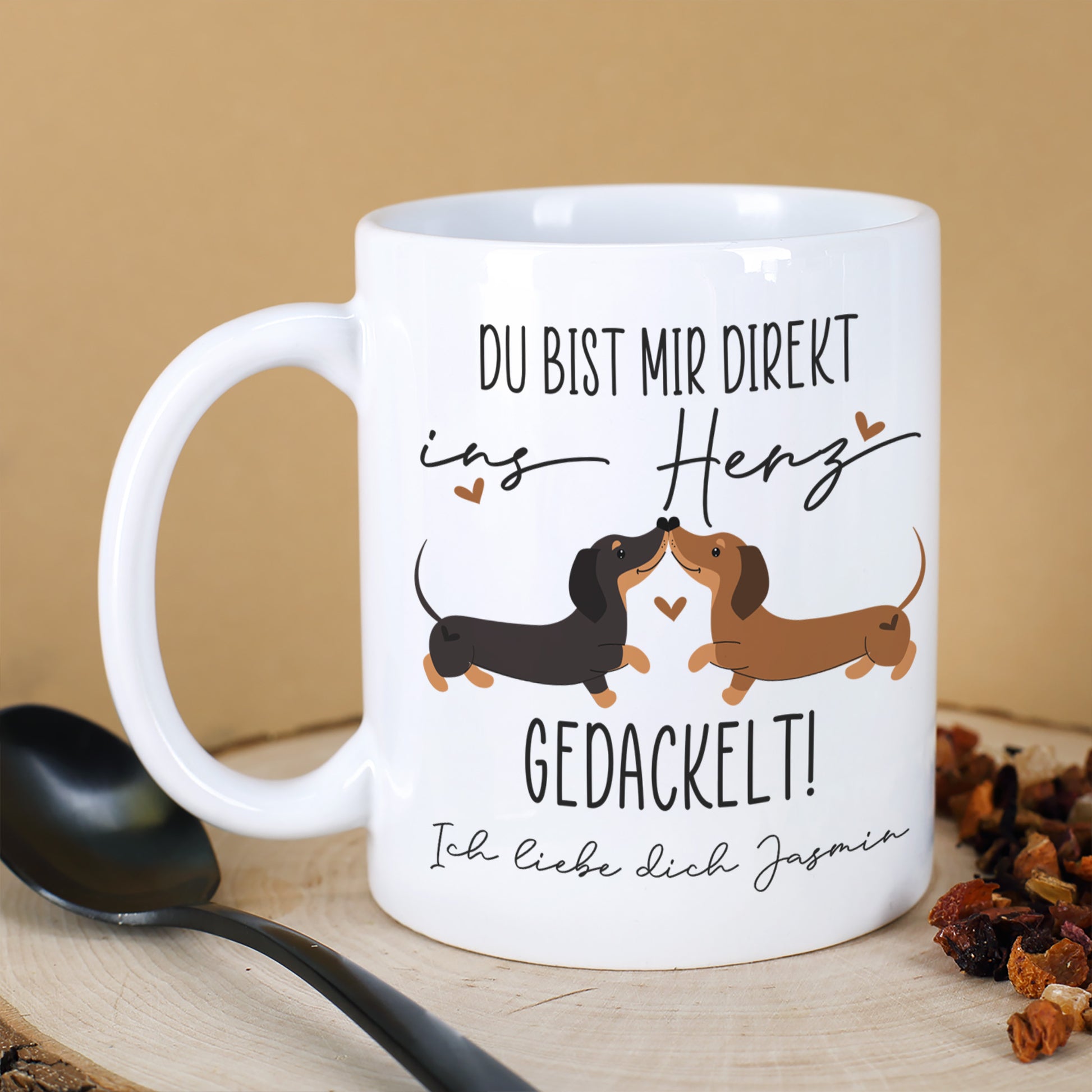 Personalisierte weiße Keramiktasse mit Dackel-Motiv und Spruch „Du bist mir direkt ins Herz gedackelt!“ auf Holzbrett, daneben Löffel und Tee