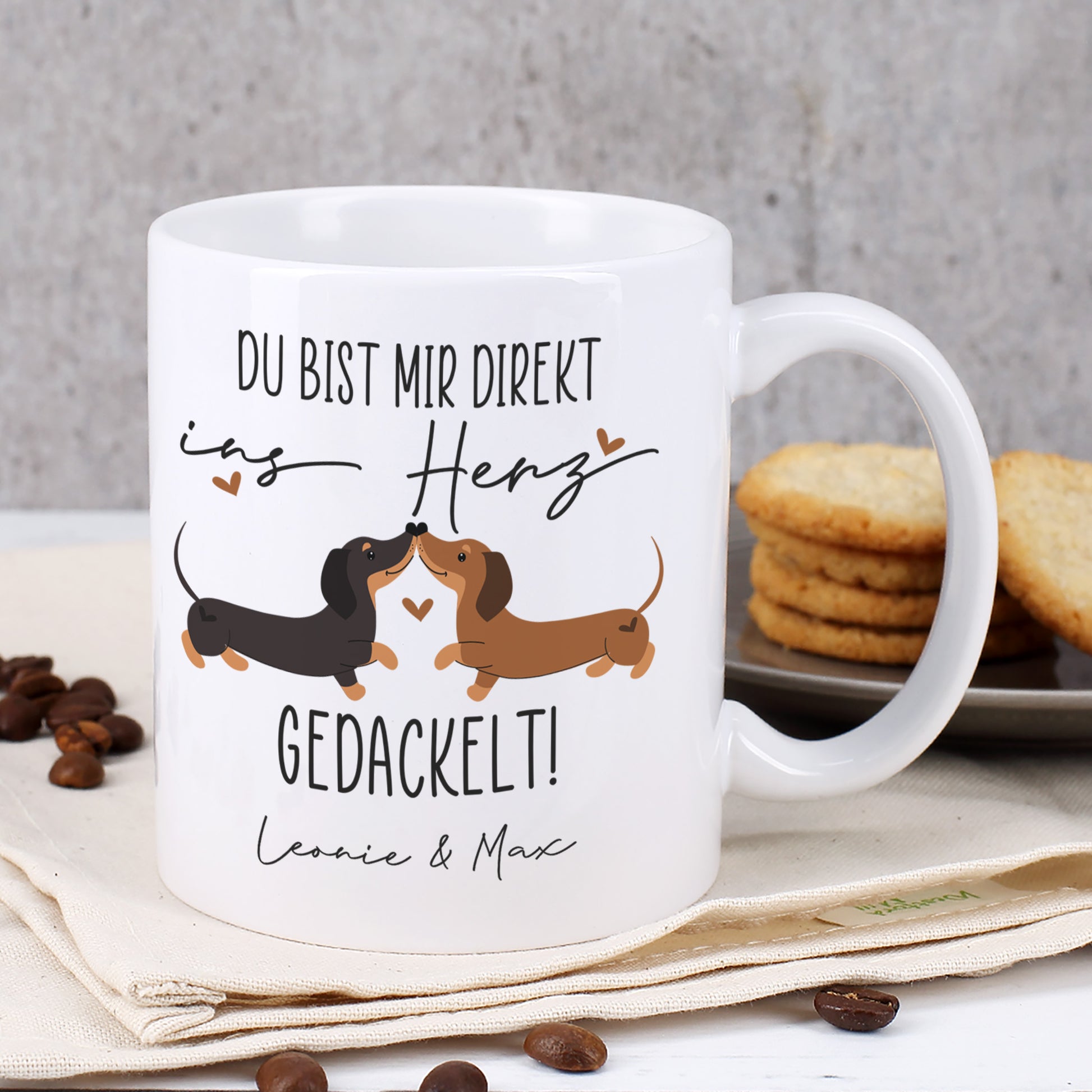 Weiße Tasse mit Dackel-Liebesmotiv und Spruch „Du bist mir direkt ins Herz gedackelt!“ vor Keksdose und Keksen, personalisiert mit Namen