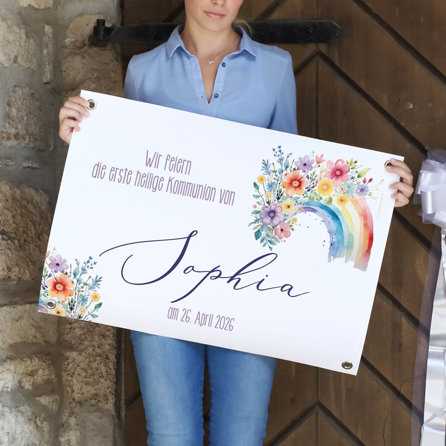 Frau hält ein personalisiertes Banner zur Kommunion in den Händen. Darauf steht: „Wir feiern die erste heilige Kommunion von Sophia – am 26. April 2026“. Das Banner zeigt einen Regenbogen aus Aquarellfarben, umgeben von einem floralen Blumenmotiv in fröhlichen Farben. Die Ecken des Banners sind mit Metallösen versehen.