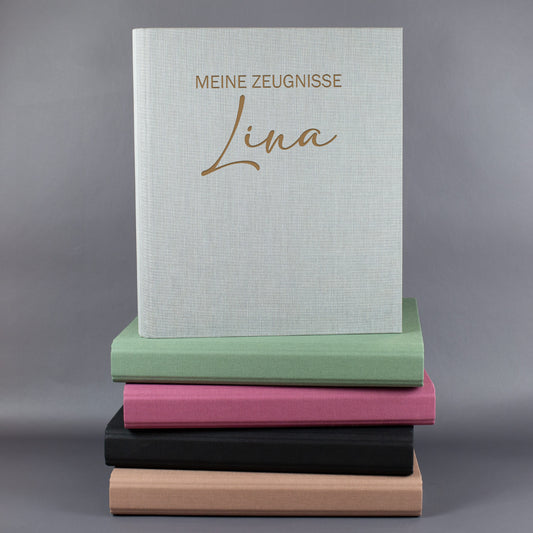 Zeugnismappe aus Leinen in Hellgrau mit Gravur „Meine Zeugnisse“ und Name Lina auf einem Stapel farbiger Mappen.