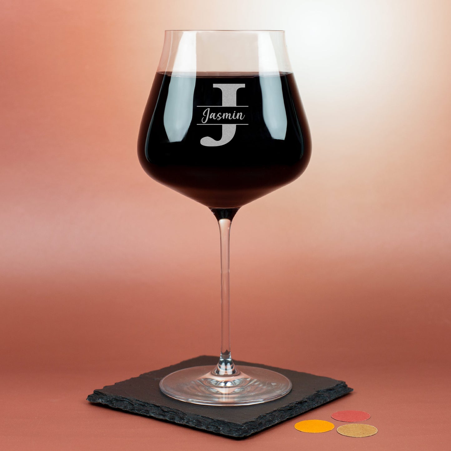 Personalisiertes Leonardo Burgunderglas mit Gravur des Buchstabens J und Name Jasmin, gefüllt mit Rotwein auf rotem Hintergrund.