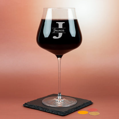 Personalisiertes Leonardo Burgunderglas mit Gravur des Buchstabens J und Name Jasmin, gefüllt mit Rotwein auf rotem Hintergrund.