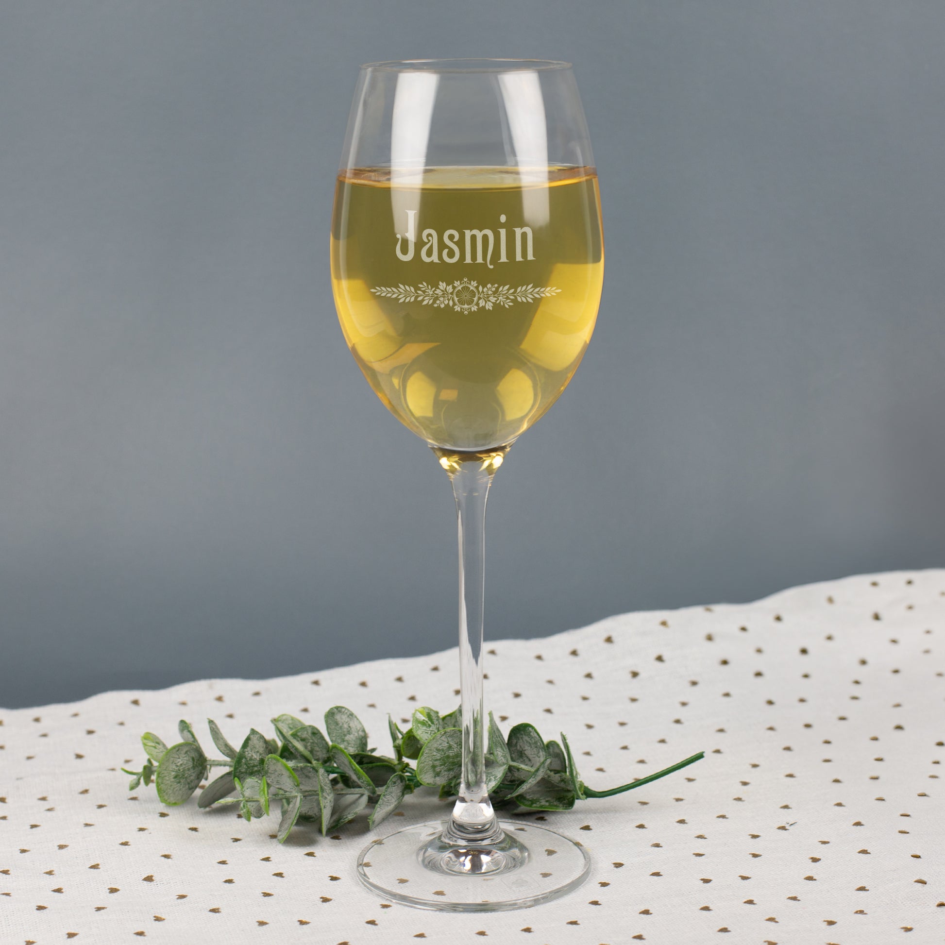 Leonardo Weißweinglas mit Gravur Jasmin und floralem Blätter-Ornament, gefüllt mit Weißwein, auf grauem Hintergrund