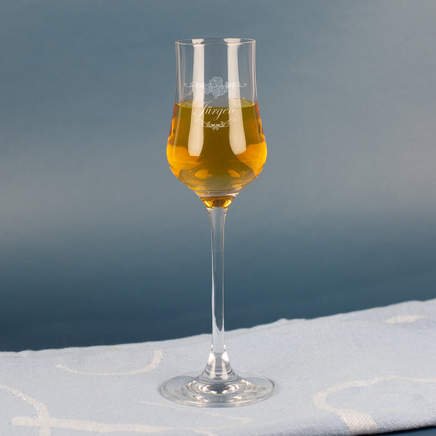 Leonardo Grappaglas mit Gravur „Jürgen“ – gefüllt mit goldfarbenem Grappa, auf blauem Hintergrund.
