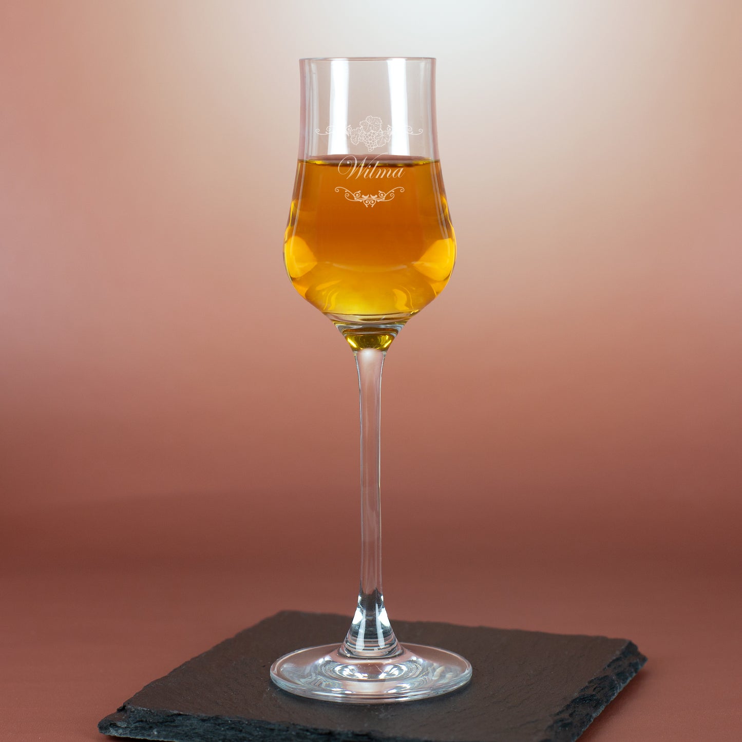 Personalisiertes Grappaglas mit Gravur „Wilma“ – stilvoll auf Schieferplatte vor rötlichem Hintergrund.
