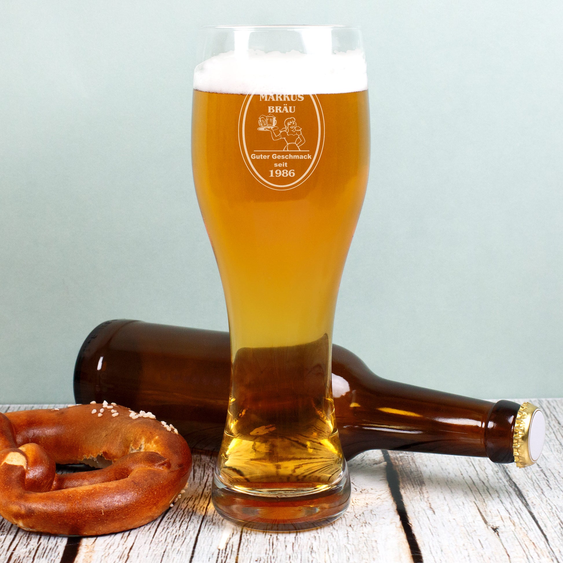 Personalisierbares Weizenglas mit Gravur-Logo „Bräu“, Name und Jahr, gefüllt mit Bier, neben Brezel und Flasche