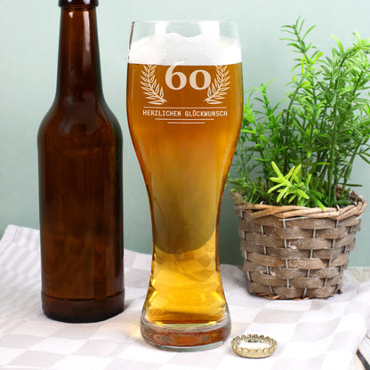 Ein gefülltes Weizenglas mit goldgelbem Bier und Schaumkrone steht neben einer braunen Bierflasche und einem Pflanzkorb mit grünem Kraut. Auf dem Glas ist eine Gravur mit Lorbeerkranz, dem Alter 60 und dem Text „Herzlichen Glückwunsch“ zu sehen.