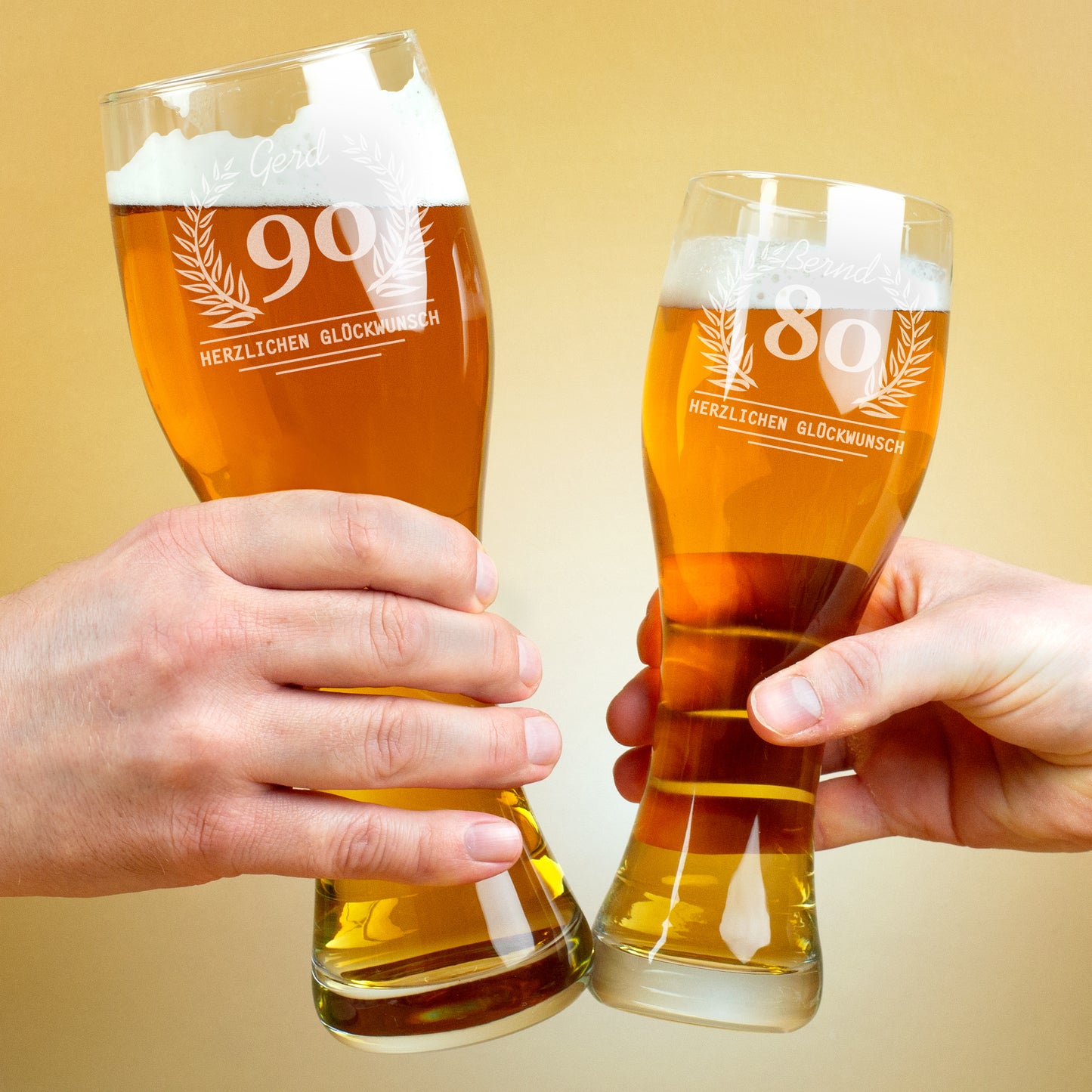 Zwei Personen stoßen mit gefüllten Weizengläsern an. Beide Gläser tragen Gravuren mit Lorbeerkranz, Alter und Namen, darunter der Schriftzug „Herzlichen Glückwunsch“.