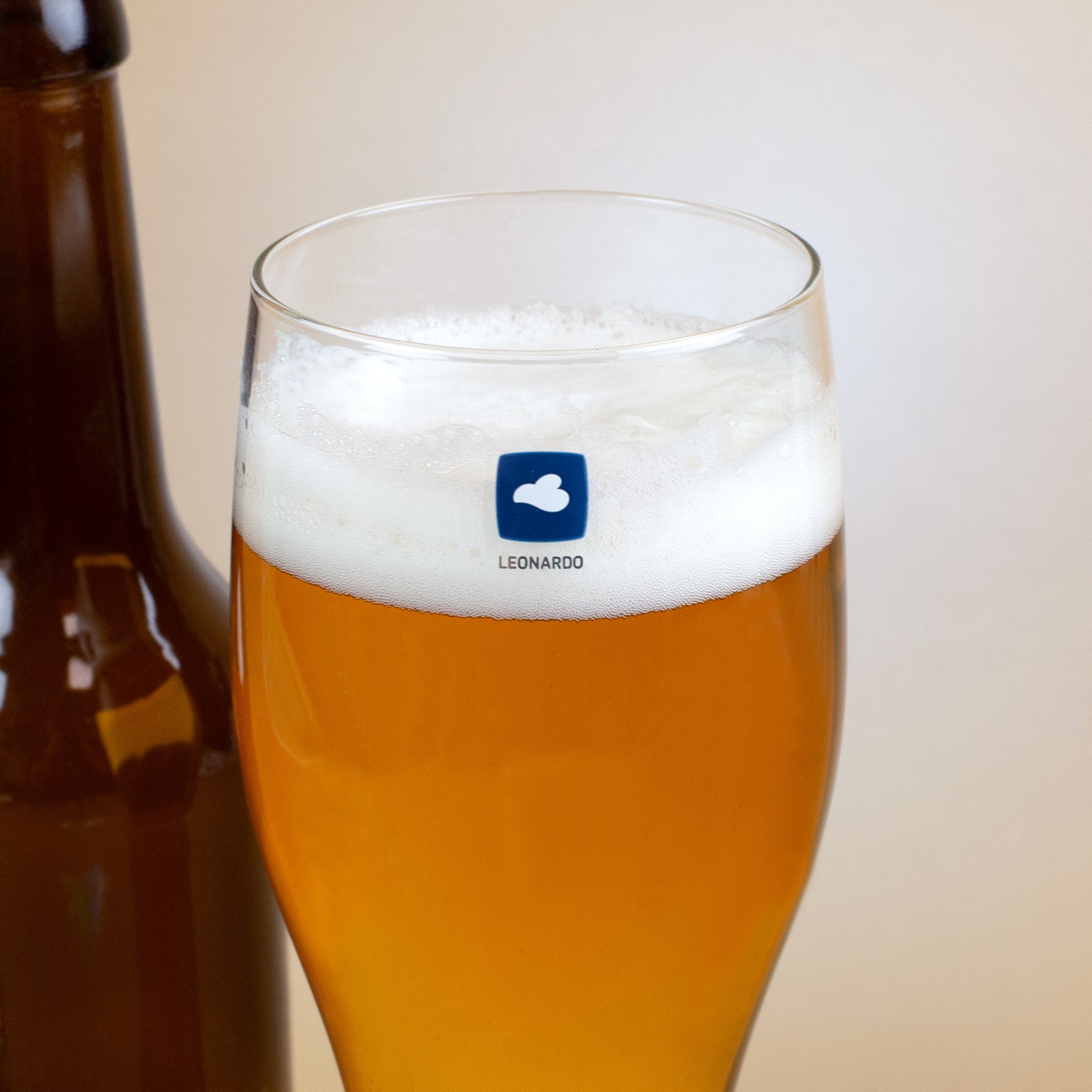 Nahaufnahme eines Leonardo-Weizenglases mit goldgelbem Bier und Schaumkrone. Auf dem Glas ist das Leonardo-Logo mit einer weißen Wolke im blauen Quadrat erkennbar, daneben eine braune Bierflasche.