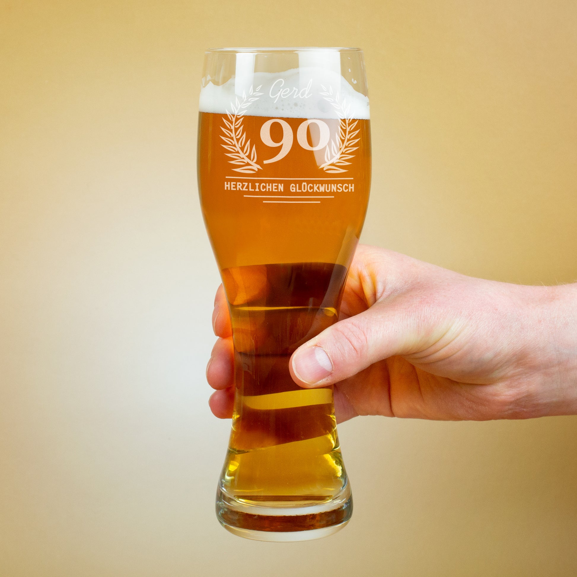 Eine Hand hält ein gefülltes Weizenglas mit Schaumkrone. Auf dem Glas ist eine Gravur mit Lorbeerkranz, der Zahl 90 und dem Schriftzug „Herzlichen Glückwunsch“ sowie einem Namen darüber zu sehen.