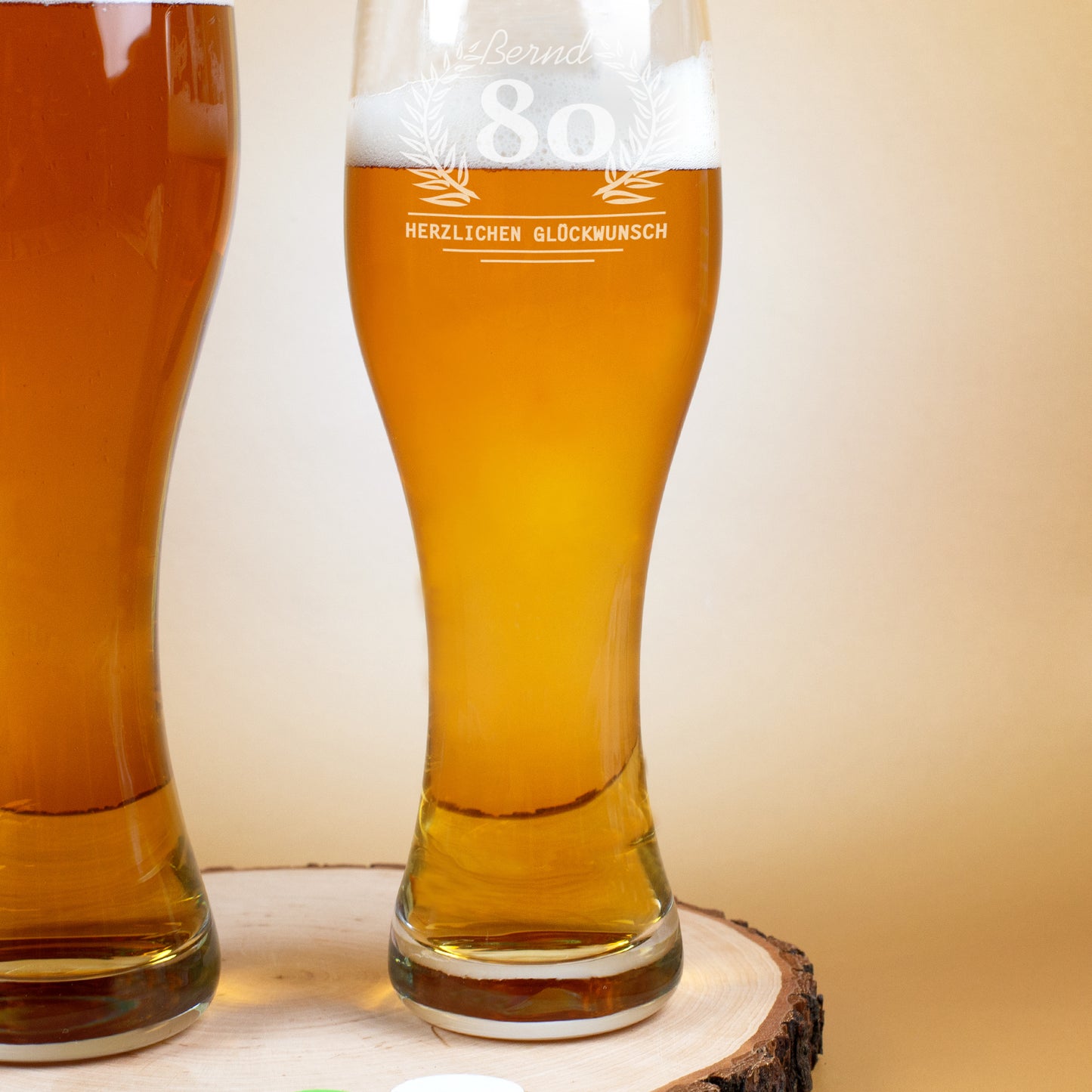 Zwei nebeneinanderstehende Weizengläser, beide gefüllt mit Bier. Auf dem rechten Glas ist eine Gravur mit Lorbeerkranz, der Zahl 80 und dem Text „Herzlichen Glückwunsch“ erkennbar.