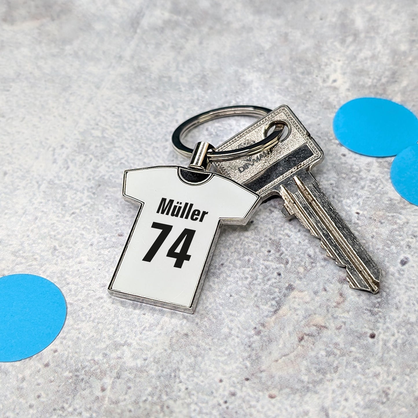 Weißer Trikot-Schlüsselanhänger mit Namen „Müller“ und Zahl „74“ an einem Schlüsselring auf hellem Untergrund