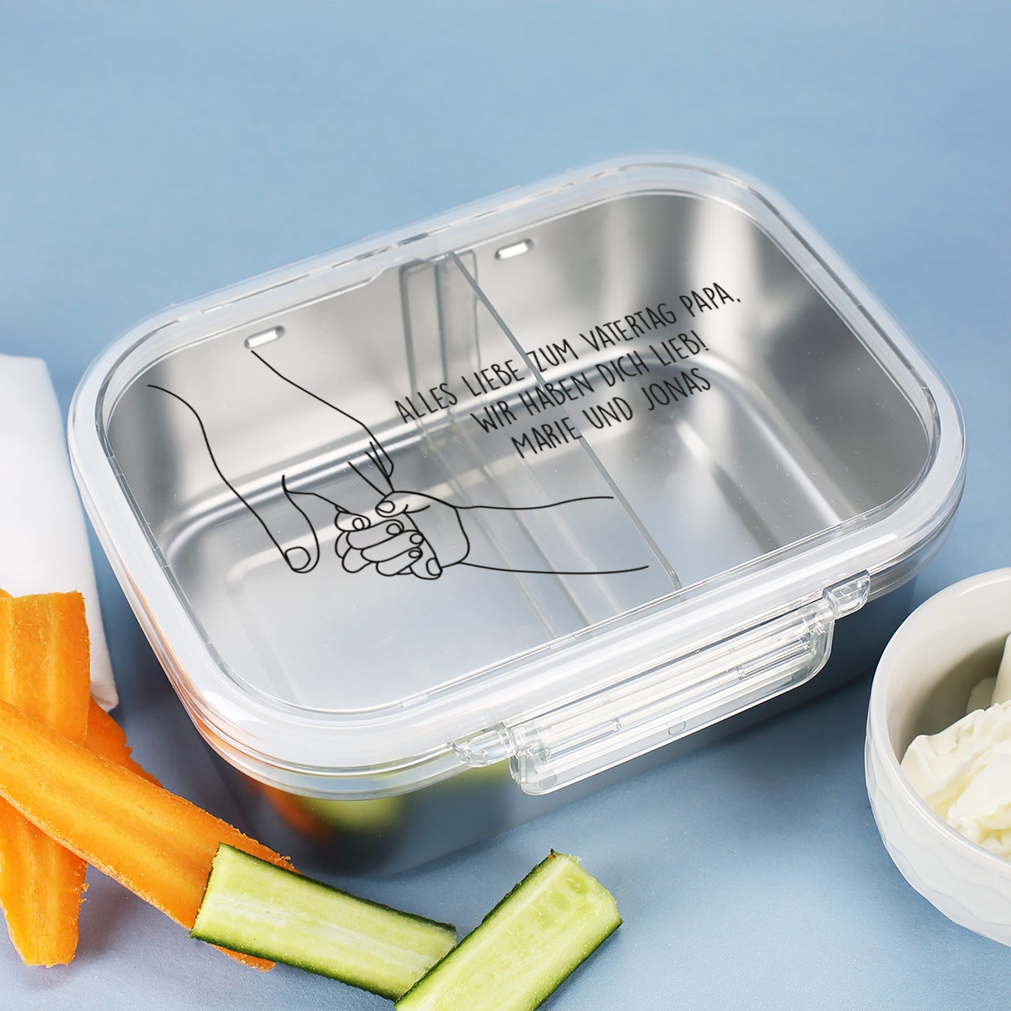 Brotdose aus Edelstahl mit Hand-in-Hand-Motiv und Text zum Vatertag, daneben Gemüsesticks