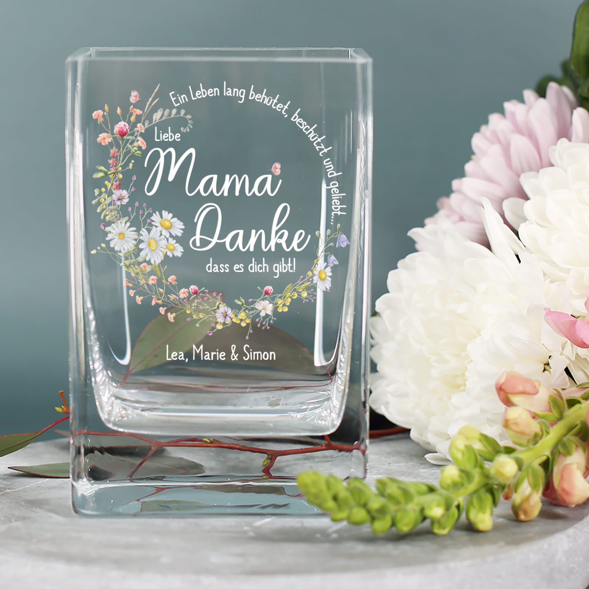 Vase mit Blumenmotiv und Aufschrift „Liebe Mama, Danke, dass es dich gibt!“ auf Tisch mit weißen Chrysanthemen, personalisiert mit „Lea, Marie & Simon“