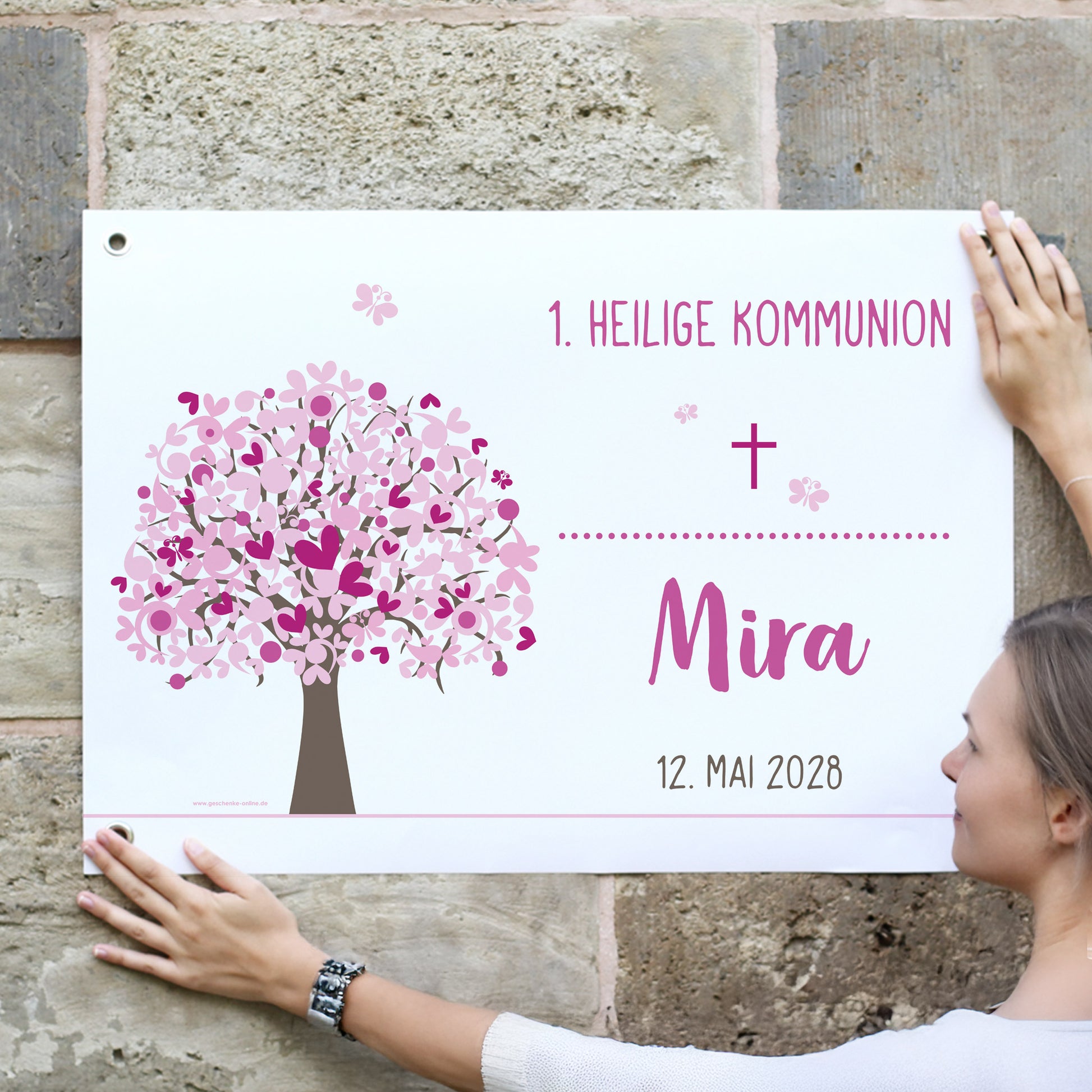 Junge Frau hängt ein rosa Lebensbaum-Banner an eine Steinmauer. Text: „1. Heilige Kommunion – Mira – 12. Mai 2028“. Blütenbaum in Rosa mit christlichen Symbolen und Namen personalisiert.