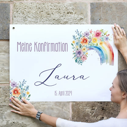 Junge Frau hängt ein personalisiertes Banner an eine Natursteinwand. Text: „Meine Konfirmation – Laura – 15. April 2029“. Das Banner ist mit einem bunten Aquarell-Regenbogen und floralen Blumenillustrationen geschmückt. Die Ecken sind mit stabilen Metallösen versehen.
